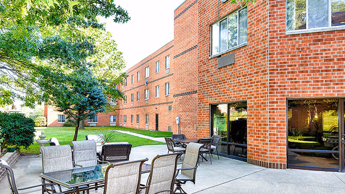 Outdoor patio with glass-top tables and chairs next to a red brick multi-story building and landscaped lawn.