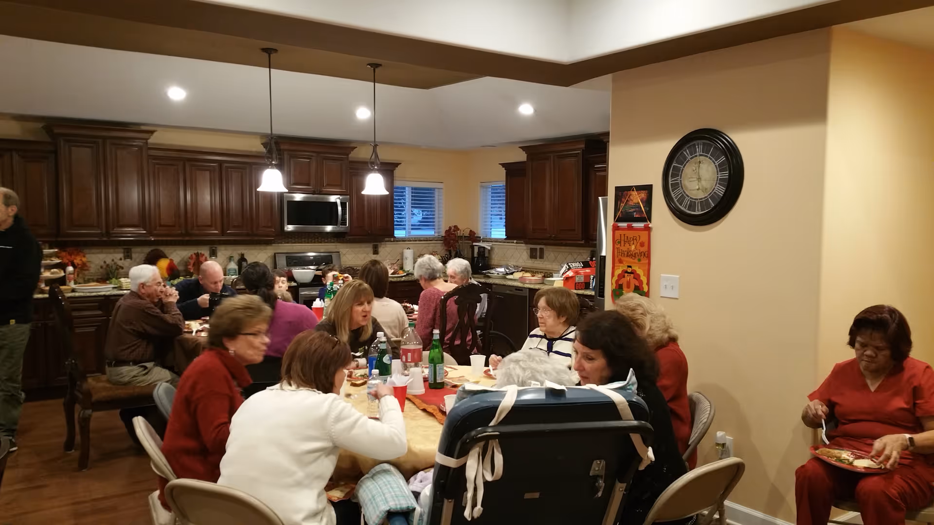 A group of elderly people and caregivers sitting around a large dining table in a kitchen area, eating and socializing. The kitchen has dark wooden cabinets, stainless steel appliances, and warm lighting. A clock and a Thanksgiving decoration are visible on the wall.