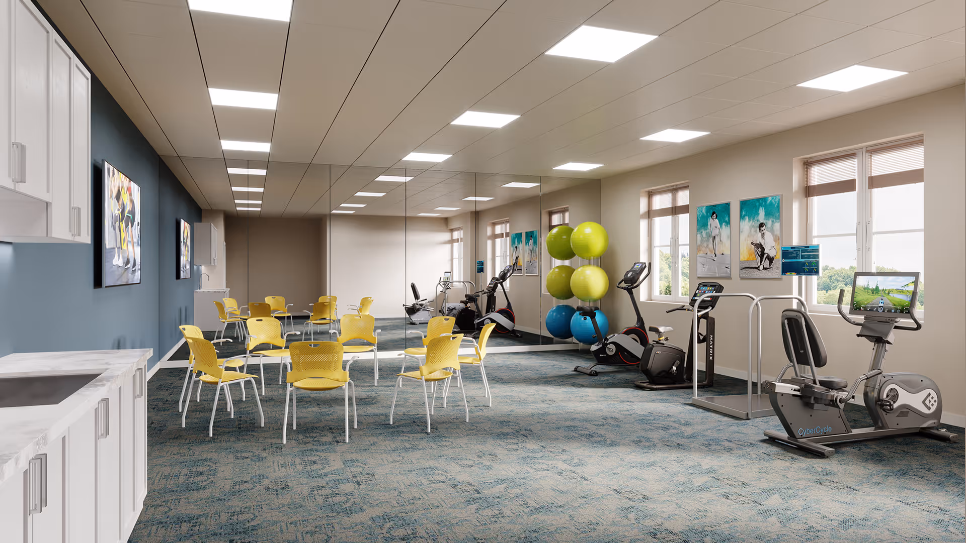 Bright multi-purpose fitness room with exercise machines, stacked stability balls, yellow chairs around tables, a mirrored wall and a small kitchenette.