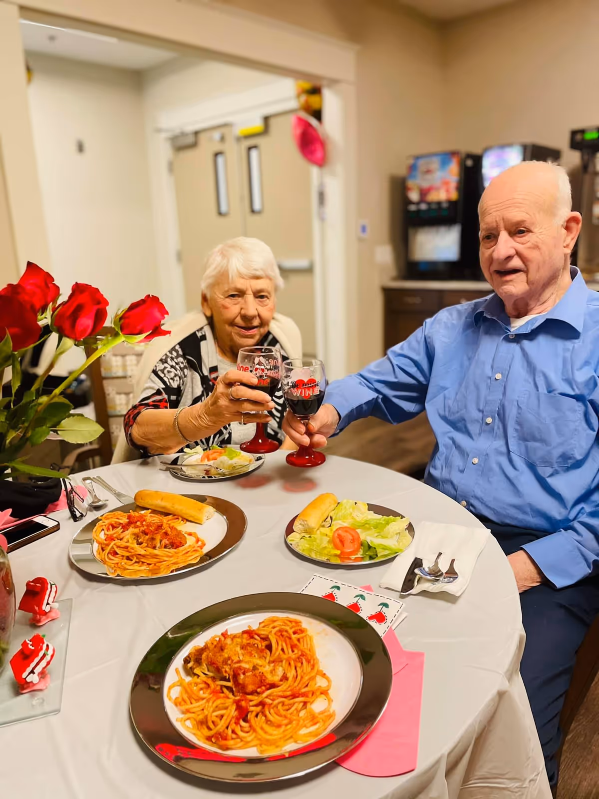 An elderly woman and man sitting at a round table in a dining area, clinking glasses of red wine. The table is set with plates of spaghetti with tomato sauce, breadsticks, and salad. There is a vase with red roses on the table and a beverage dispenser in the background.