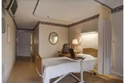 A hospital-style patient room with a single bed, a bedside table with a lamp, a chair, a wall-mounted television, and a privacy curtain partially drawn. The room has wallpaper with a decorative border near the ceiling and a round mirror on the wall.