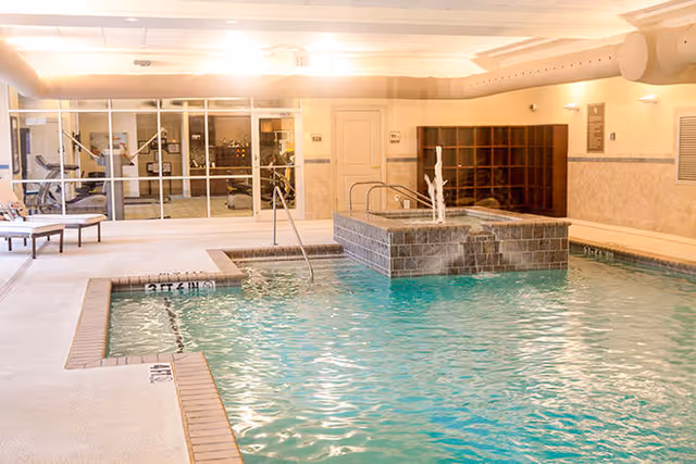 Indoor swimming pool area with clear blue water, a small raised hot tub with a water fountain feature, poolside lounge chairs, and a glass wall separating the pool from an adjacent exercise room with gym equipment.