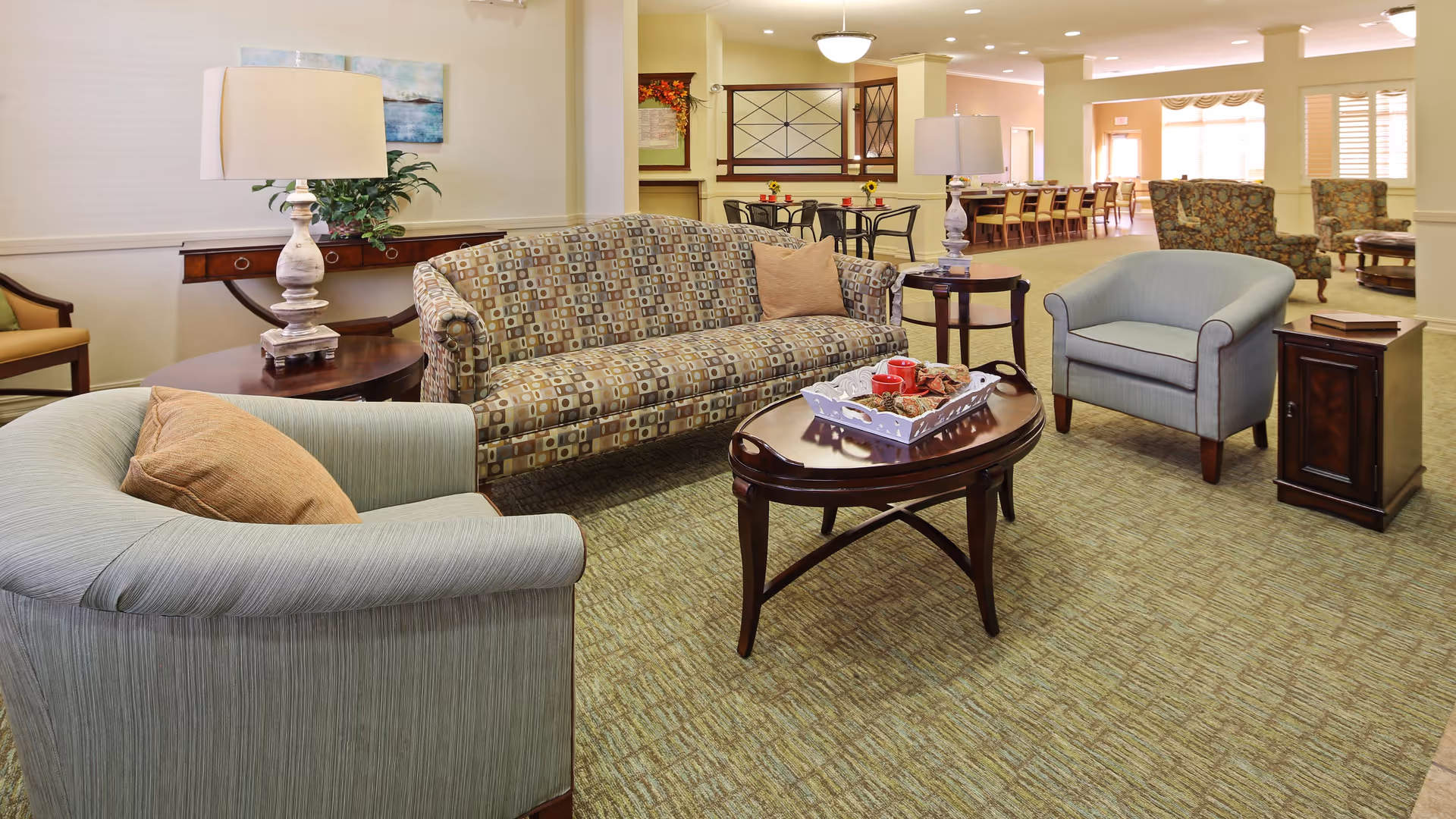 A cozy assisted living facility common area with a patterned sofa, two light blue armchairs with brown cushions, a wooden coffee table with a decorative tray, side tables with lamps, and a dining area with tables and chairs in the background.