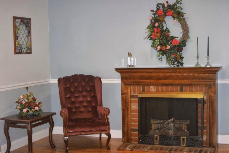 A cozy corner of a room featuring a dark red tufted armchair next to a wooden side table with a floral arrangement. On the right side, there is a brick fireplace with a wooden mantel decorated with a floral wreath and two green candles in silver candle holders.
