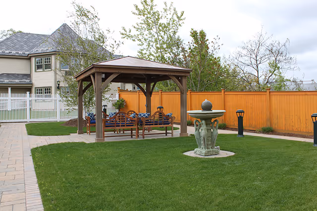 Outdoor garden area with a wooden gazebo containing benches with blue cushions, a stone fountain on a grassy lawn, surrounded by a wooden fence and a paved walkway, with a large house visible in the background.