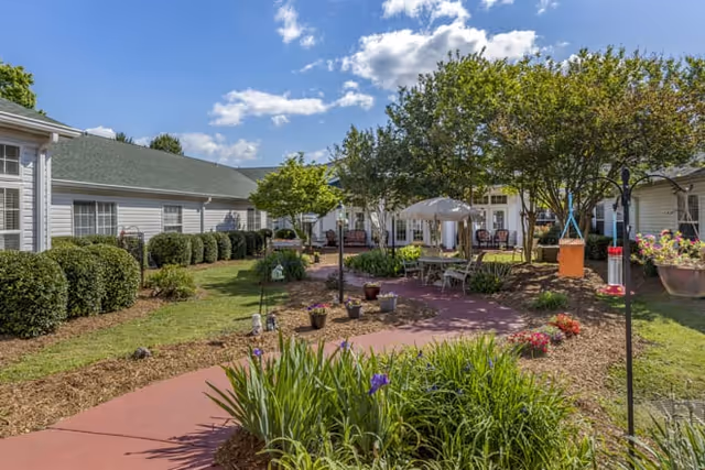 Outdoor courtyard area at Vickery Parke Assisted Living featuring a paved walkway, green bushes, flowering plants, trees, and a seating area with tables and umbrellas under a partly cloudy blue sky.