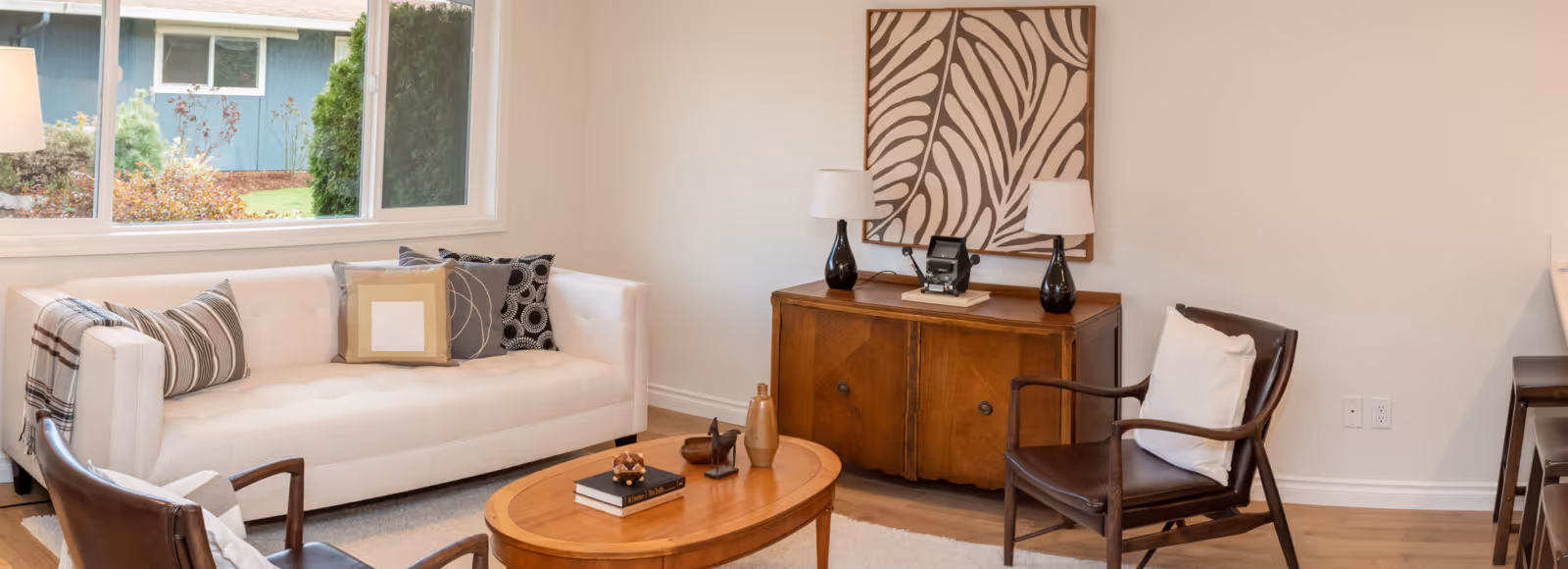 A cozy living room with a white sofa adorned with multiple decorative pillows and a plaid throw blanket. Two dark wooden armchairs with white cushions face a wooden oval coffee table that holds books and decorative items. Against the wall is a wooden sideboard with two black lamps and a zebra-patterned artwork above it. A large window shows a view of greenery outside.