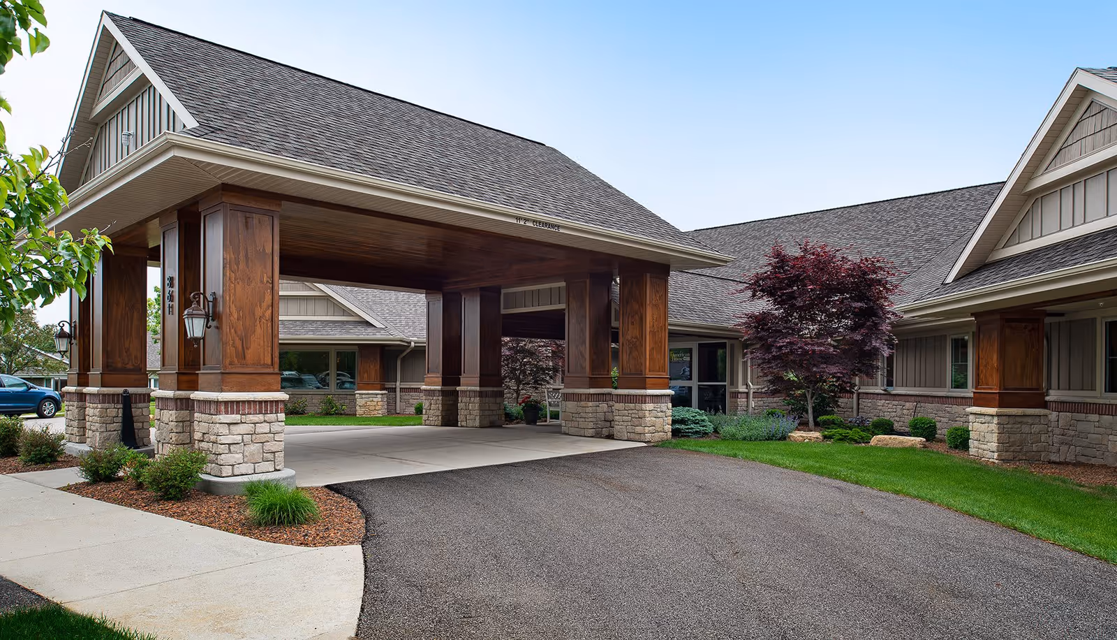 Covered porte-cochere entrance of a senior living building with stone pillars, wooden beams, and landscaped grounds.