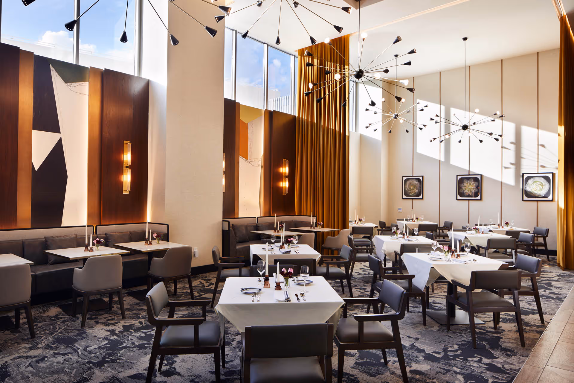 Bright, modern dining room with multiple tables set with white tablecloths, upholstered chairs, tall windows and contemporary chandeliers.