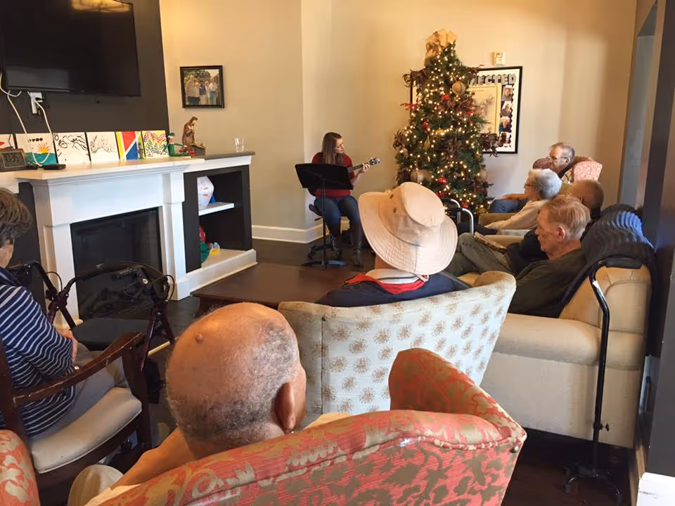 A group of elderly people seated in a living room area watching a woman playing guitar near a decorated Christmas tree. The room has a fireplace, a TV mounted on the wall, and various chairs and sofas arranged for the audience.