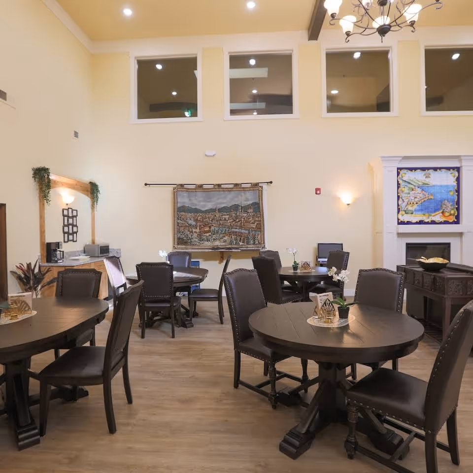 A well-lit common area with several round wooden tables and chairs arranged for seating. The room features high ceilings with multiple windows near the top, a chandelier, wall sconces, a tapestry hanging on the wall, and a fireplace with decorative artwork above it. There is also a small counter area with a coffee maker and some plants for decoration.