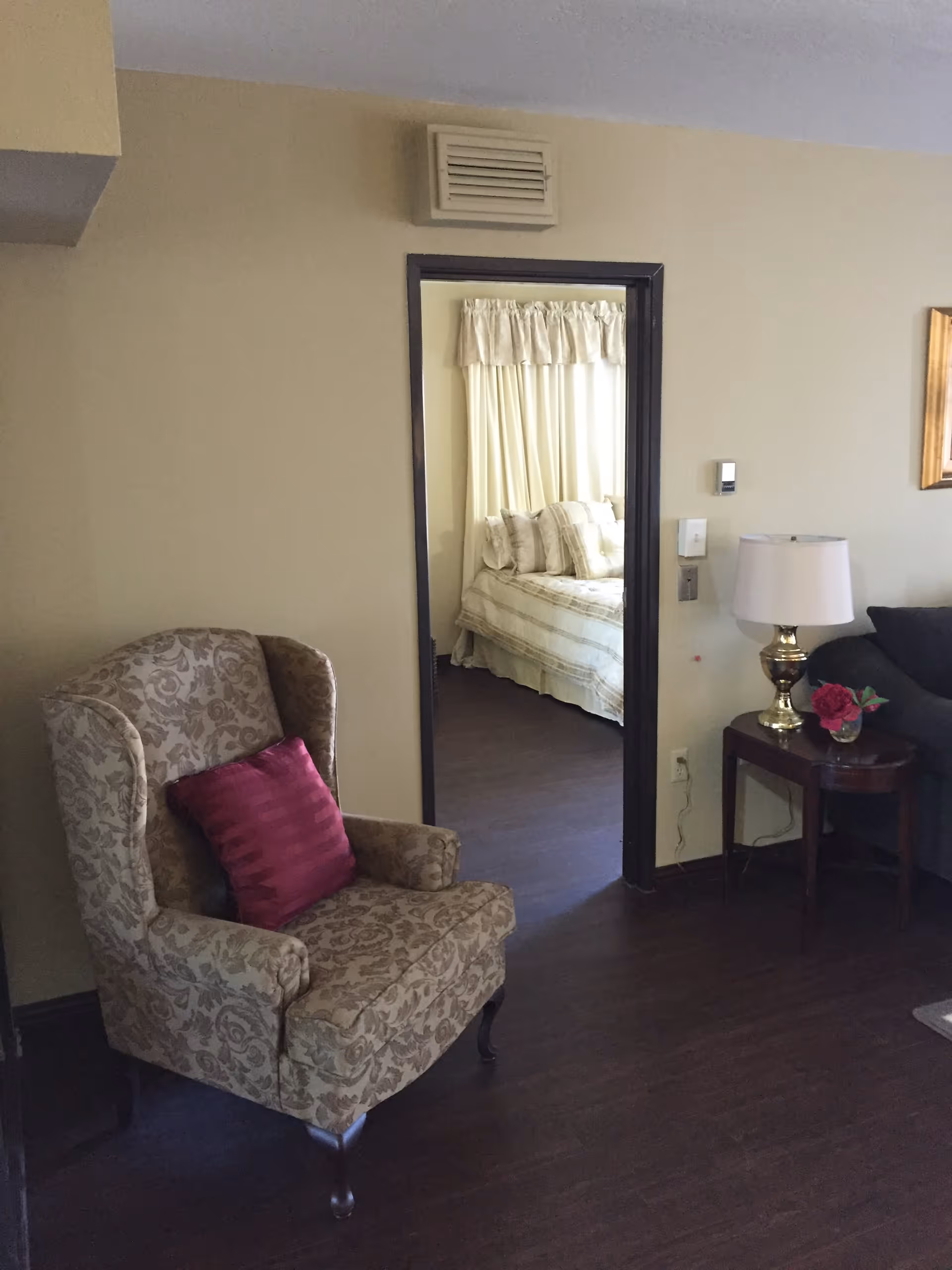 A living room with an upholstered armchair and side table with a lamp, looking through an open doorway into a bedroom.