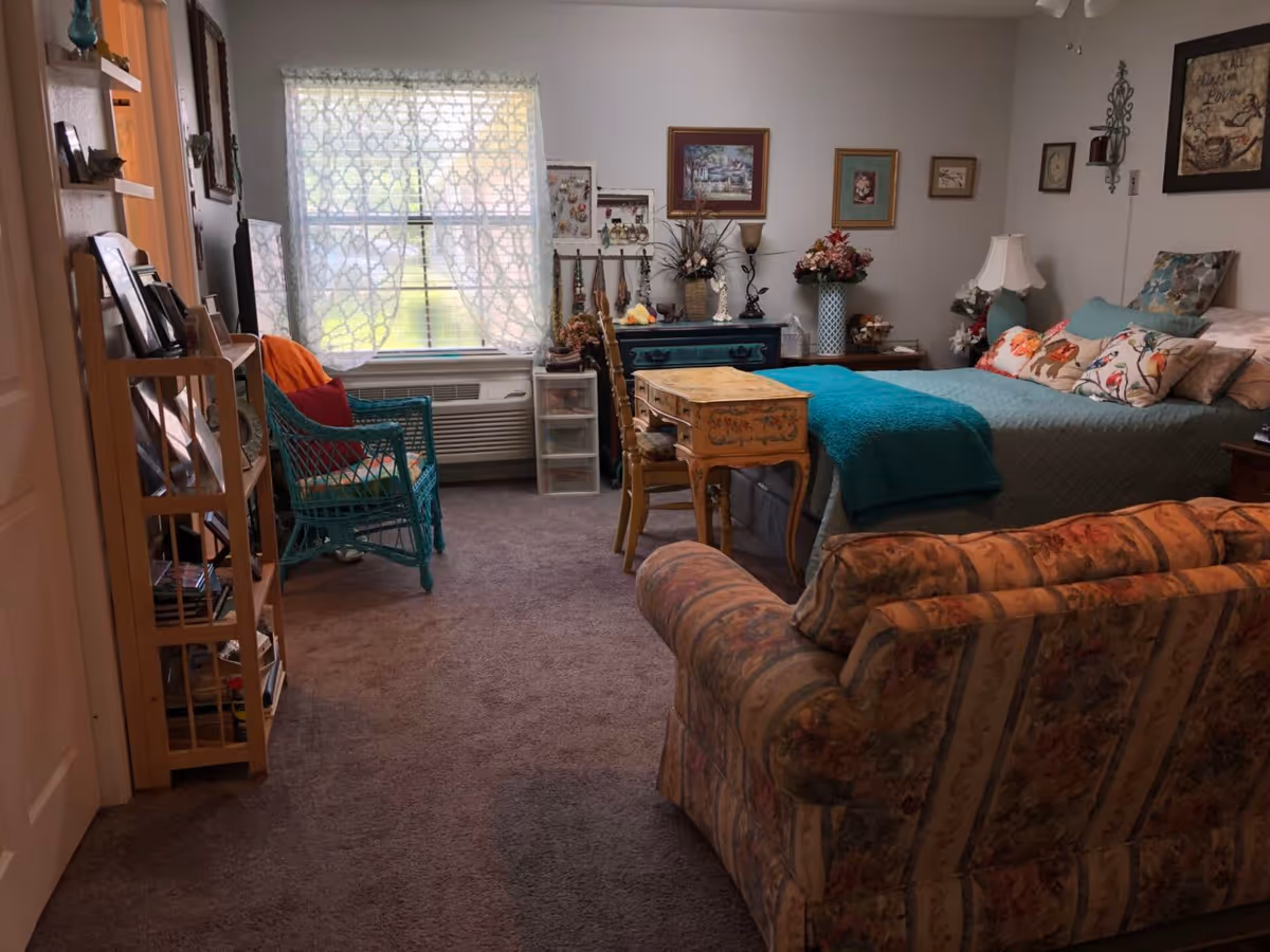 A cozy bedroom with a bed covered in teal bedding and multiple decorative pillows. There is a floral-patterned couch in the foreground, a small wooden desk with a chair at the foot of the bed, and a teal wicker chair near a window with patterned curtains. The walls are decorated with framed pictures and floral arrangements, and there is a wooden shelving unit with various items on the left side.