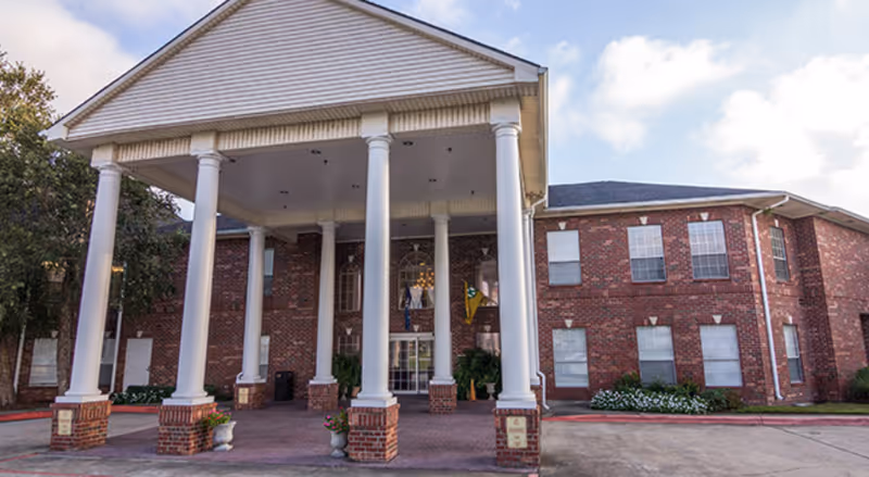 Front exterior view of a two-story brick building with large white columns supporting a covered entrance. The building has multiple windows and is surrounded by some greenery and a paved driveway under a partly cloudy sky.