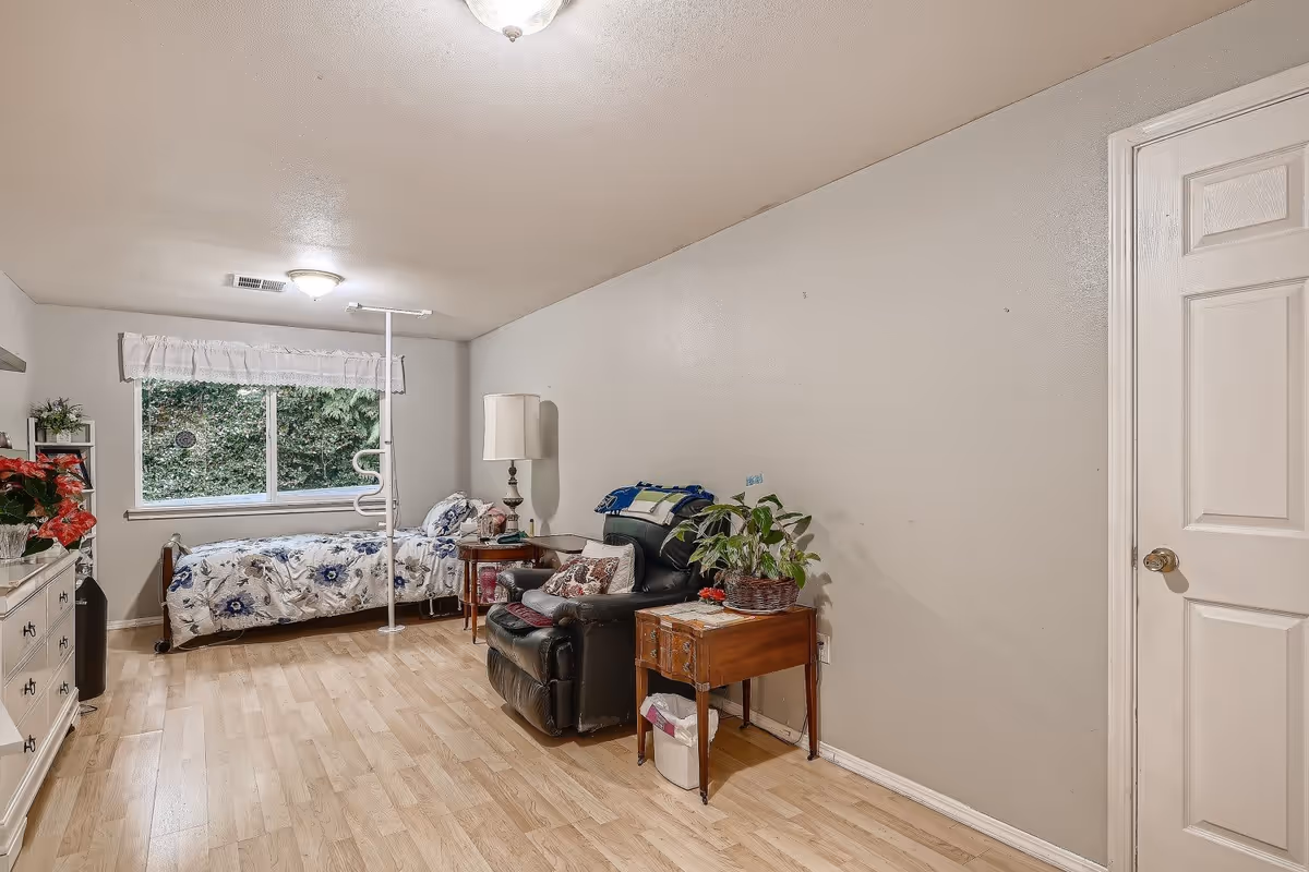 A simple bedroom with light wood flooring, a single bed with floral bedding near a window with a white valance, a black recliner chair with a small wooden side table holding a plant, a white dresser with red flowers on top, and a closed white door on the right wall.