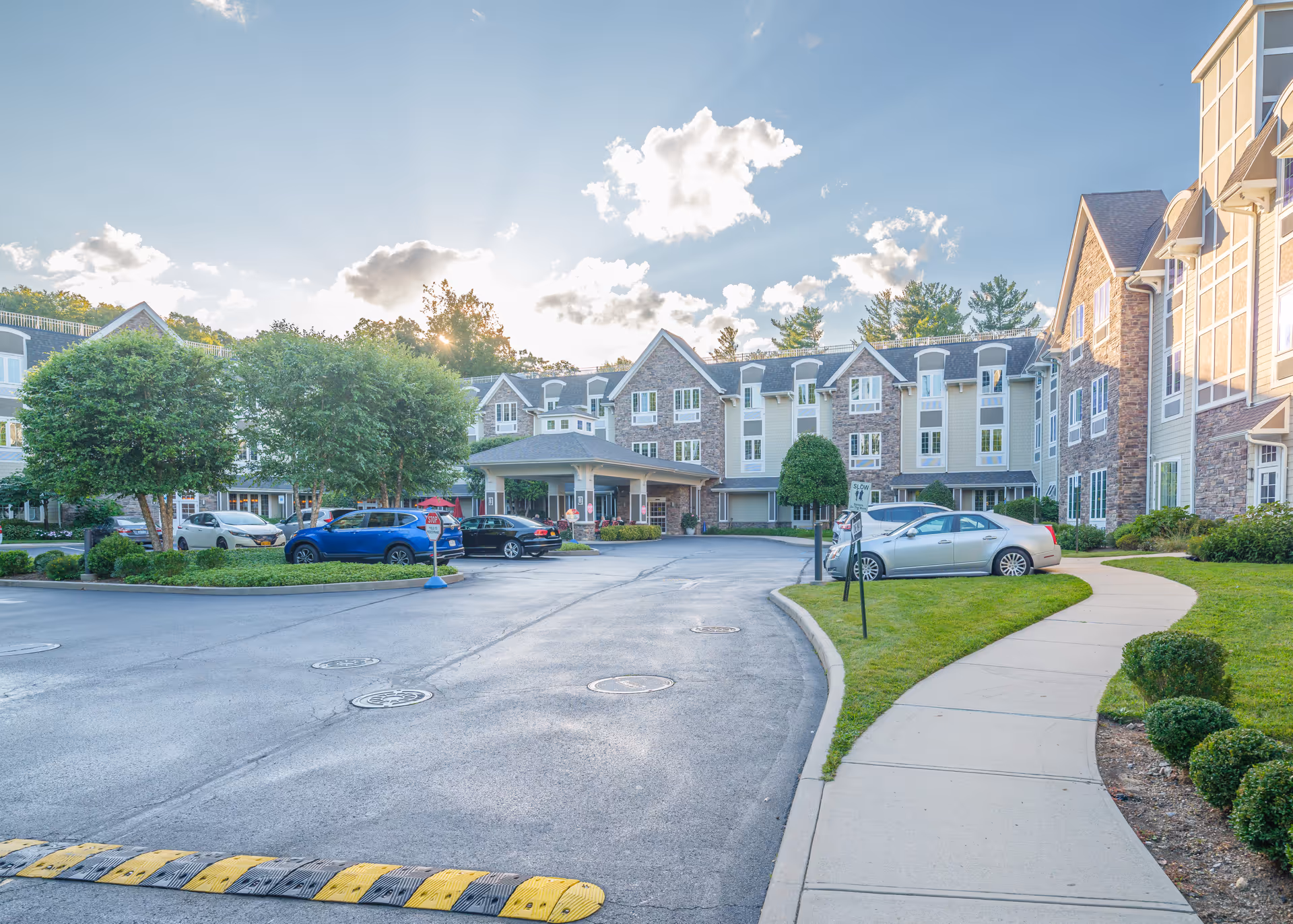 Exterior view of The Ambassador of Scarsdale senior living facility showing a large multi-story building with stone and beige siding, multiple windows, and a covered entrance. There is a paved driveway with parked cars, a sidewalk, manicured bushes, and trees under a partly cloudy sky with sunlight.