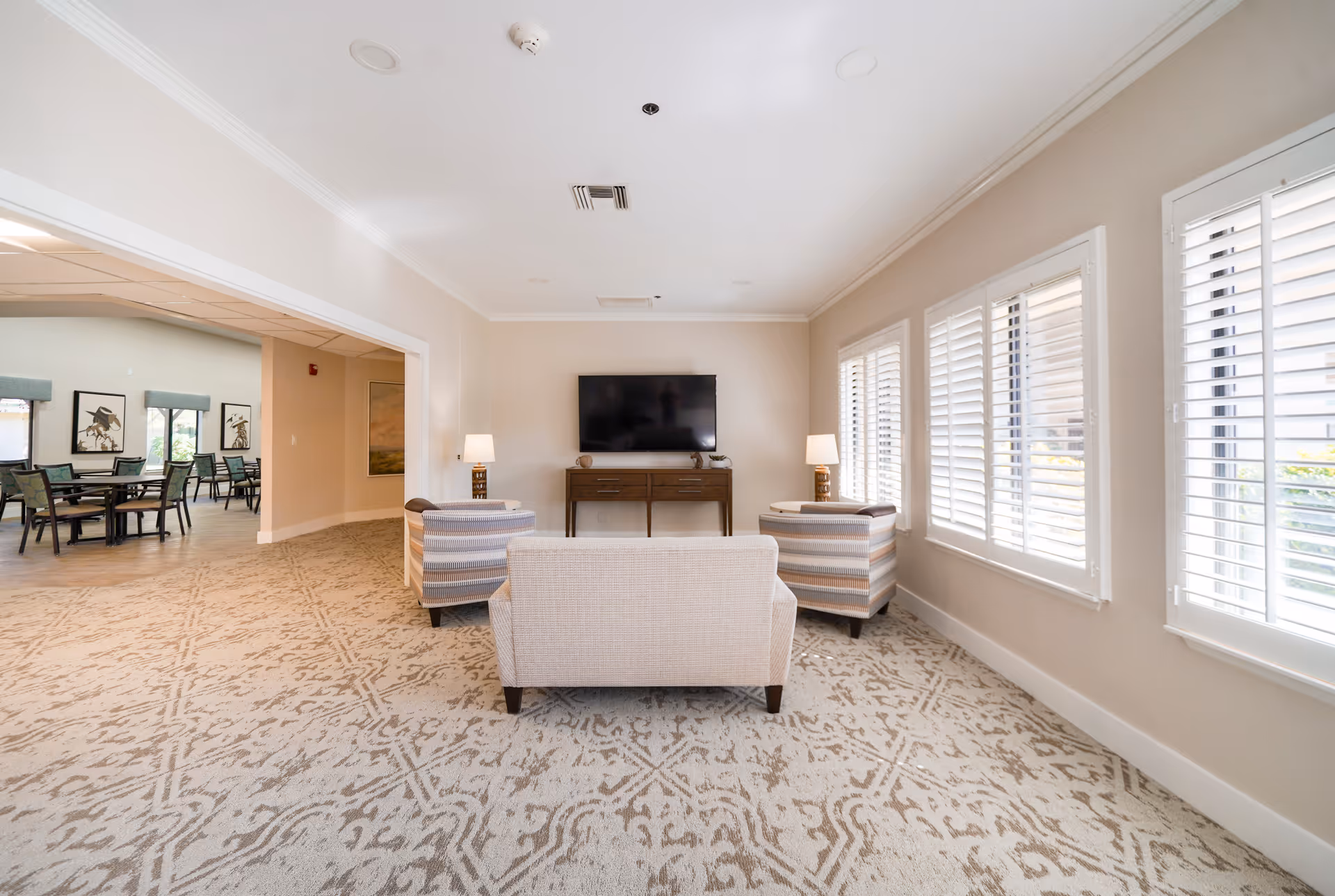A bright and spacious living room area in a senior living facility with a beige couch and two striped armchairs facing a wall-mounted flat-screen TV. The room has large windows with white plantation shutters allowing natural light to fill the space. Two table lamps are placed on either side of the TV on a wooden console. Adjacent to the living room is a dining area with multiple tables and chairs.
