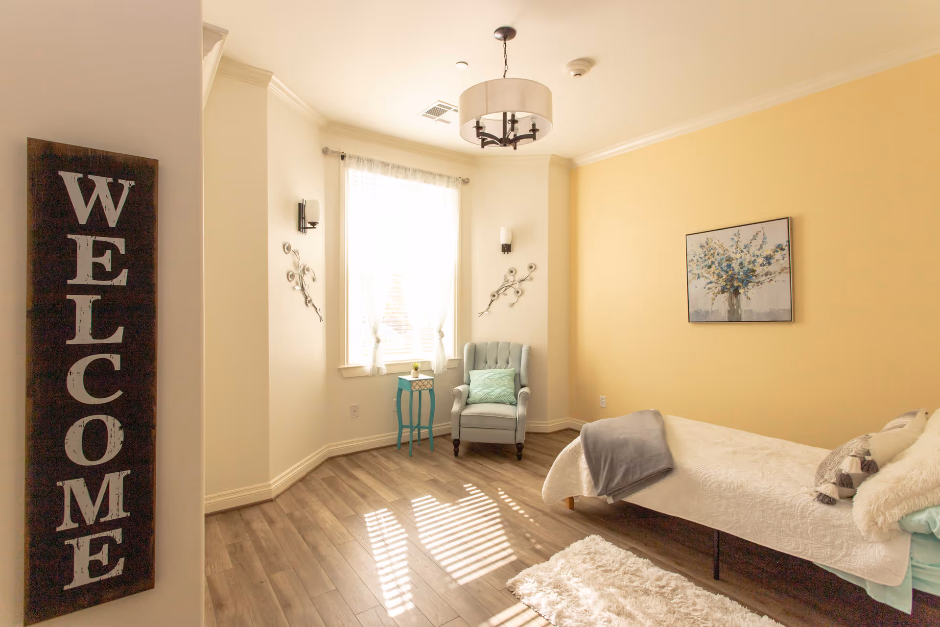 A cozy bedroom with a single bed covered in white bedding and a gray throw blanket. There is a light blue armchair with a green pillow next to a window with sheer white curtains. A small turquoise side table with a decorative item sits beside the chair. The walls are painted light yellow and cream, with a floral painting hanging above the bed. A wooden sign with the word 'WELCOME' is mounted on the left wall. The room has wood flooring and a white rug near the bed.