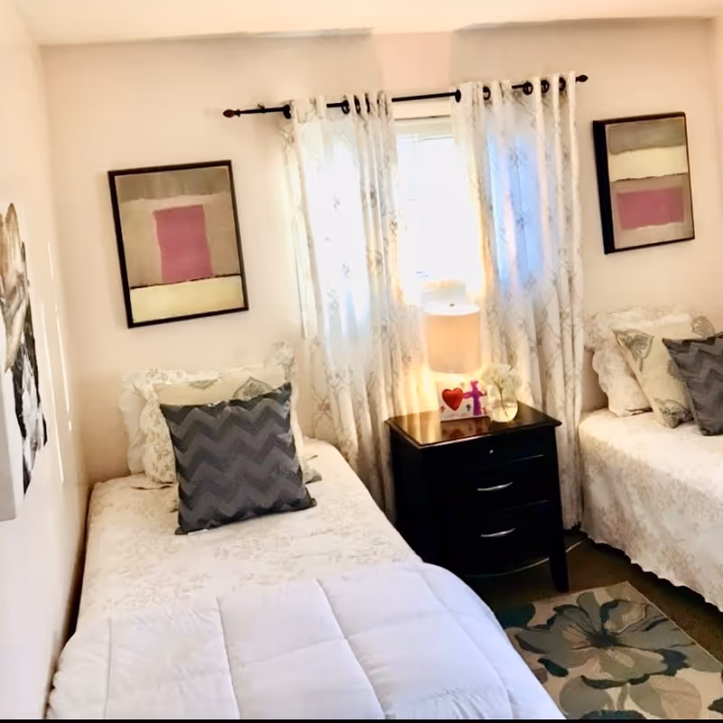 Two twin beds with patterned pillows and a nightstand holding a lamp between them in a softly lit bedroom with curtains and framed artwork.