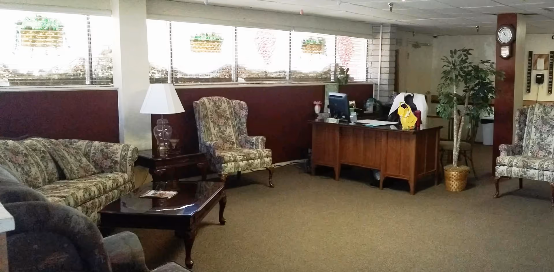 A cozy interior room with floral upholstered armchairs and a sofa, a wooden coffee table, a side table with a lamp, a wooden desk with a computer and office chair, a potted plant, and a wall clock.