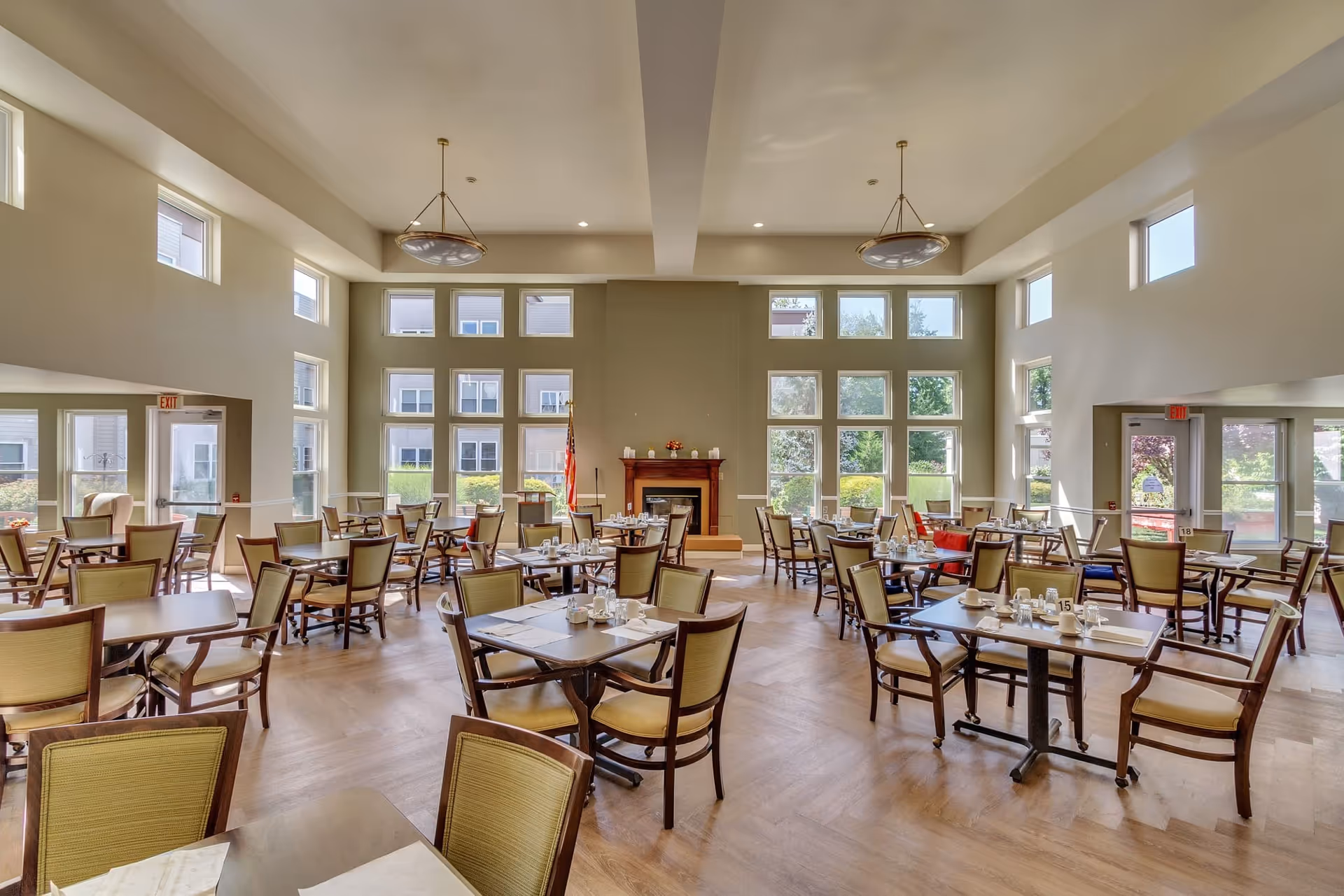 A spacious dining room with multiple tables and chairs arranged neatly. The room has large windows letting in natural light, a fireplace with decorative items on the mantel, and two modern ceiling light fixtures. The floor is wooden, and the room has a calm, inviting atmosphere.