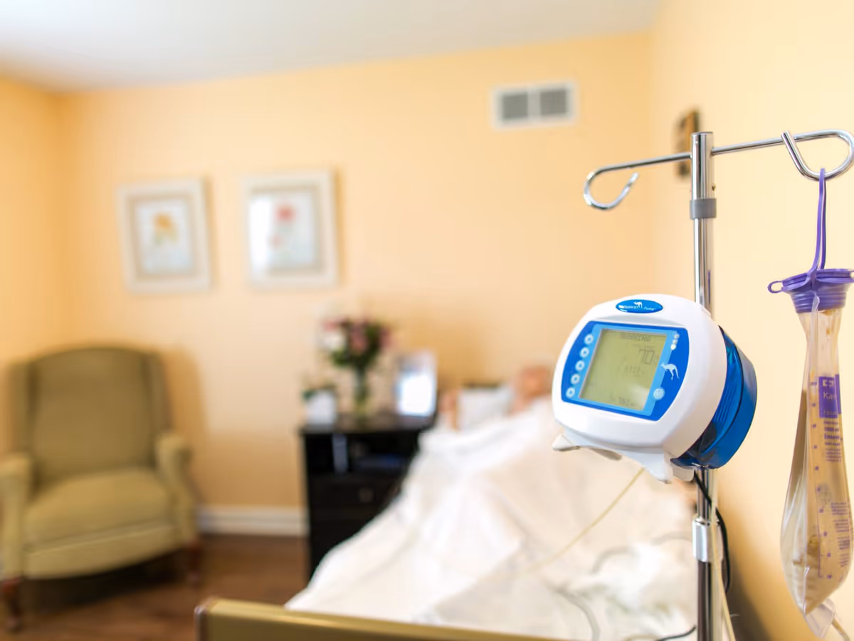 A medical infusion pump attached to an IV pole in the foreground with a blurred background showing a patient lying in bed covered with a white blanket, a green armchair, a nightstand with flowers, and framed pictures on a beige wall.
