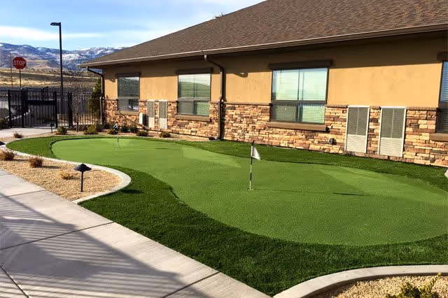 Outdoor putting green area next to a building with stone and stucco exterior walls, featuring small flags on the green and a sidewalk with landscaping around it. Mountains and a stop sign are visible in the background.