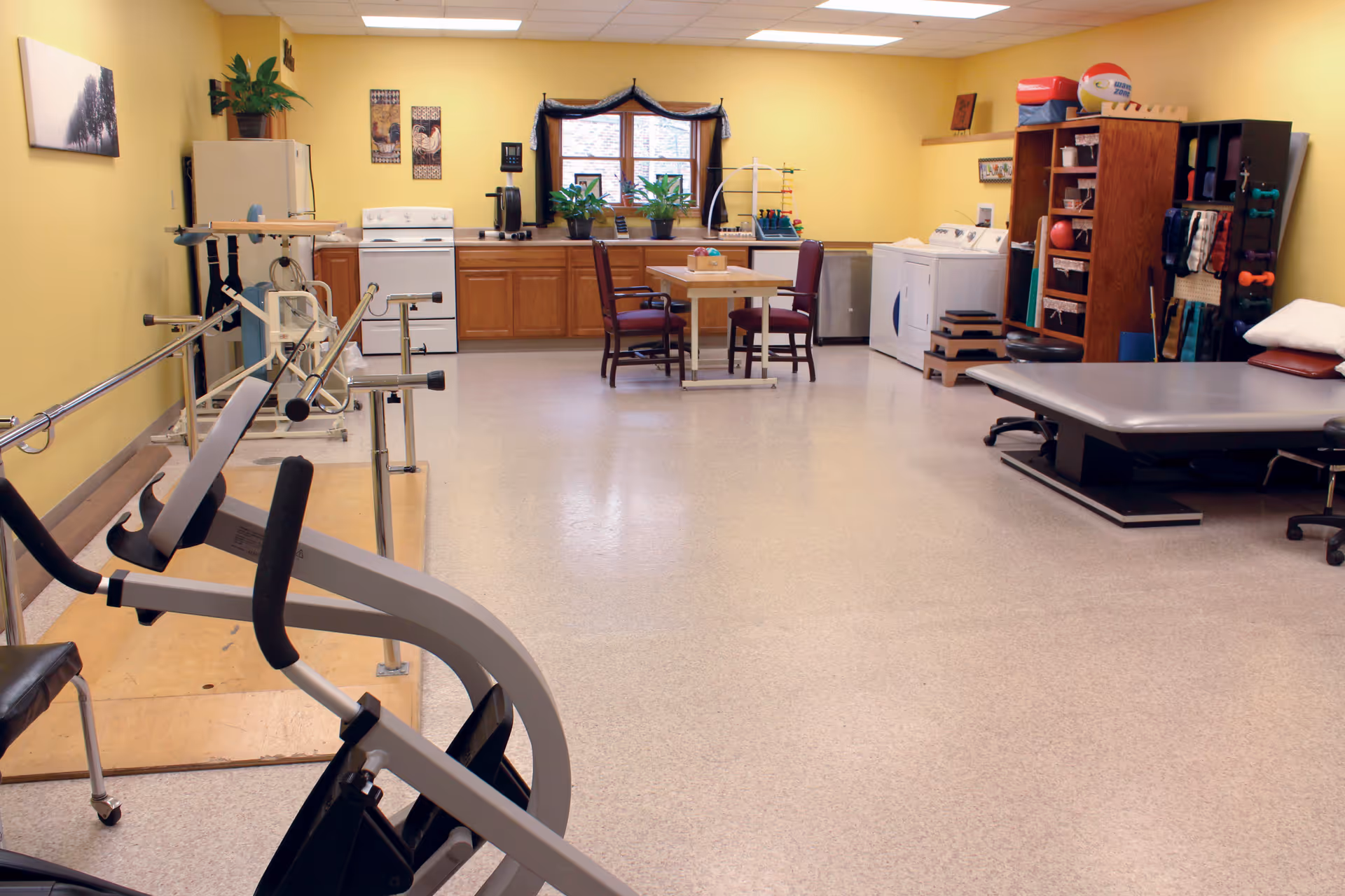 A spacious room with yellow walls featuring exercise equipment, parallel bars, a therapy table, and a small table with two chairs. The back wall has a window with plants on the counter below it, a stove, a refrigerator, and a washing machine. Shelves with various exercise and therapy tools are also visible.