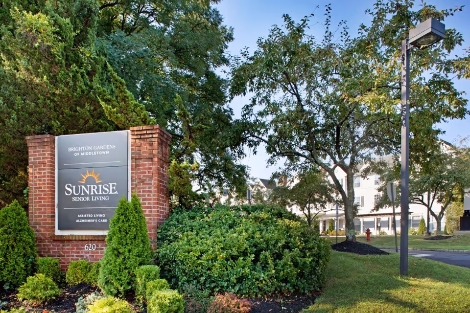 Outdoor view of Brighton Gardens of Middletown senior living facility sign made of brick with the Sunrise Senior Living logo, surrounded by green bushes and trees, with a building and streetlamp visible in the background.