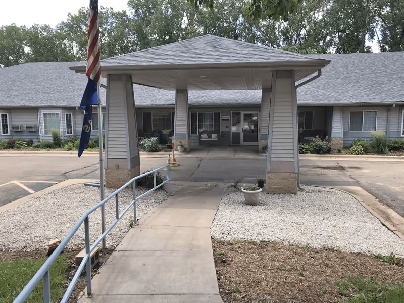 Entrance to a single-story building with a covered drop-off area supported by four pillars. There is a concrete walkway with a metal handrail leading to the entrance. Two flags are displayed on a flagpole to the left of the walkway. The building has multiple windows and a gray shingled roof, surrounded by some landscaping with gravel and plants.