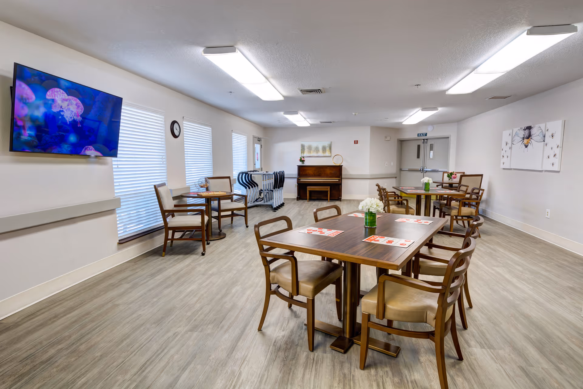 Bright communal dining/activity room with several tables and chairs, a wall-mounted TV, piano, and large windows.