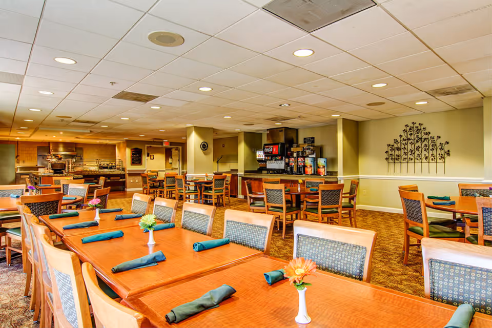 A spacious dining room in a senior living facility with multiple wooden tables and chairs arranged neatly. Each table has a small vase with a flower and green cloth napkins. In the background, there is a beverage station with soda dispensers and a buffet area with food warmers. The walls are light-colored with decorative metal wall art, and the ceiling has recessed lighting.
