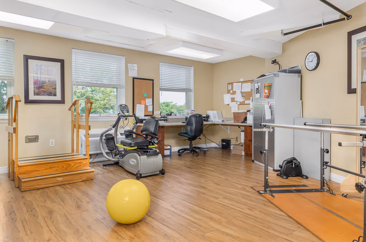 A bright therapy/exercise room with a recumbent exercise bike, parallel bars, therapy stairs, a desk area, and a yellow exercise ball on a wood floor.