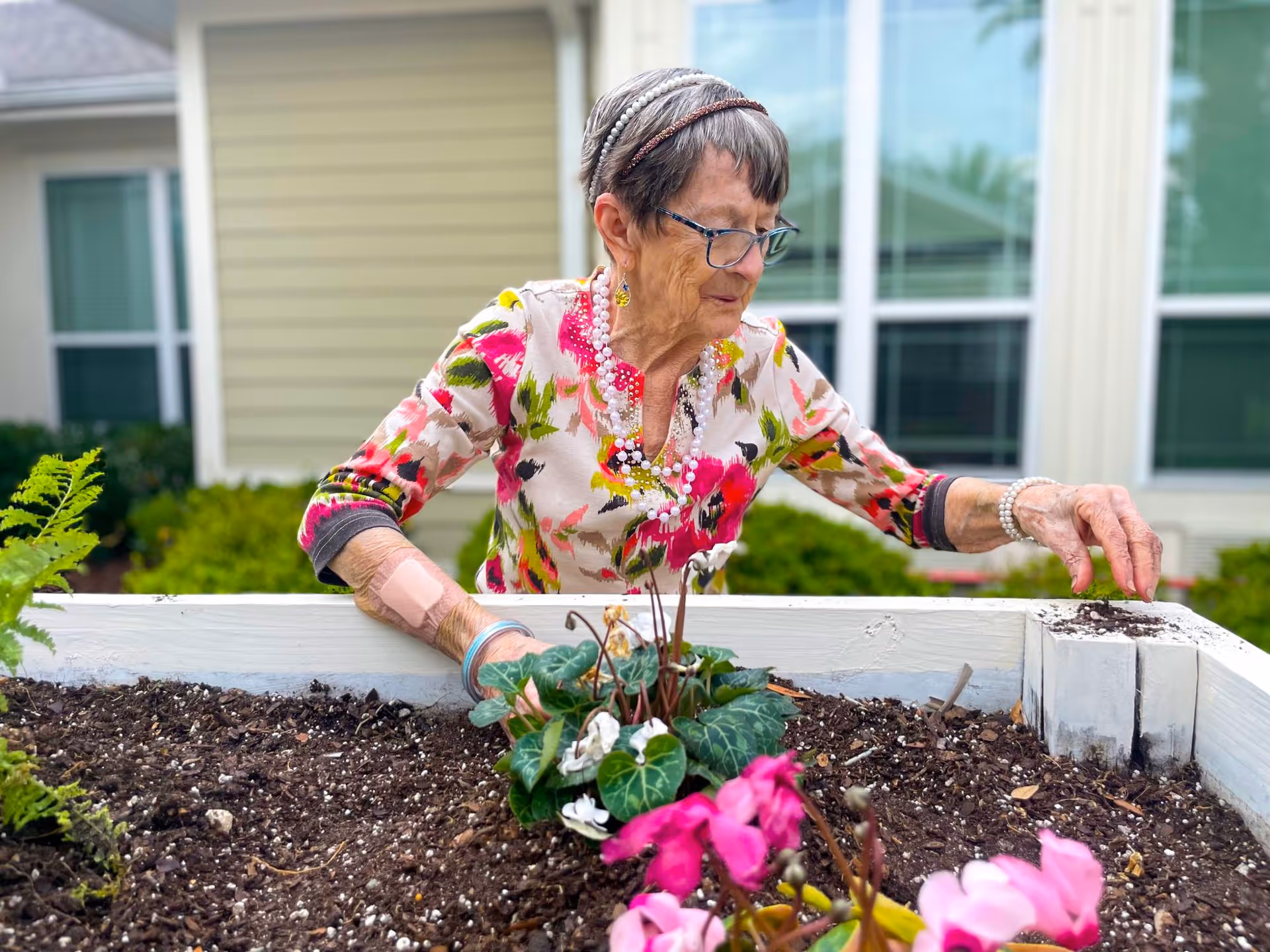 An elderly woman wearing glasses and a colorful floral blouse is gardening in a raised garden bed outside a residential building. She is tending to plants with pink flowers and appears focused on her task. The background shows windows and siding of the building.