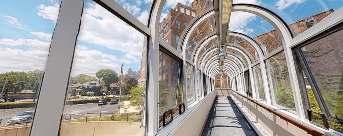 A long enclosed glass walkway with arched windows on both sides, connecting two buildings. Outside, there is a view of a parking lot, trees, and residential houses under a partly cloudy sky.