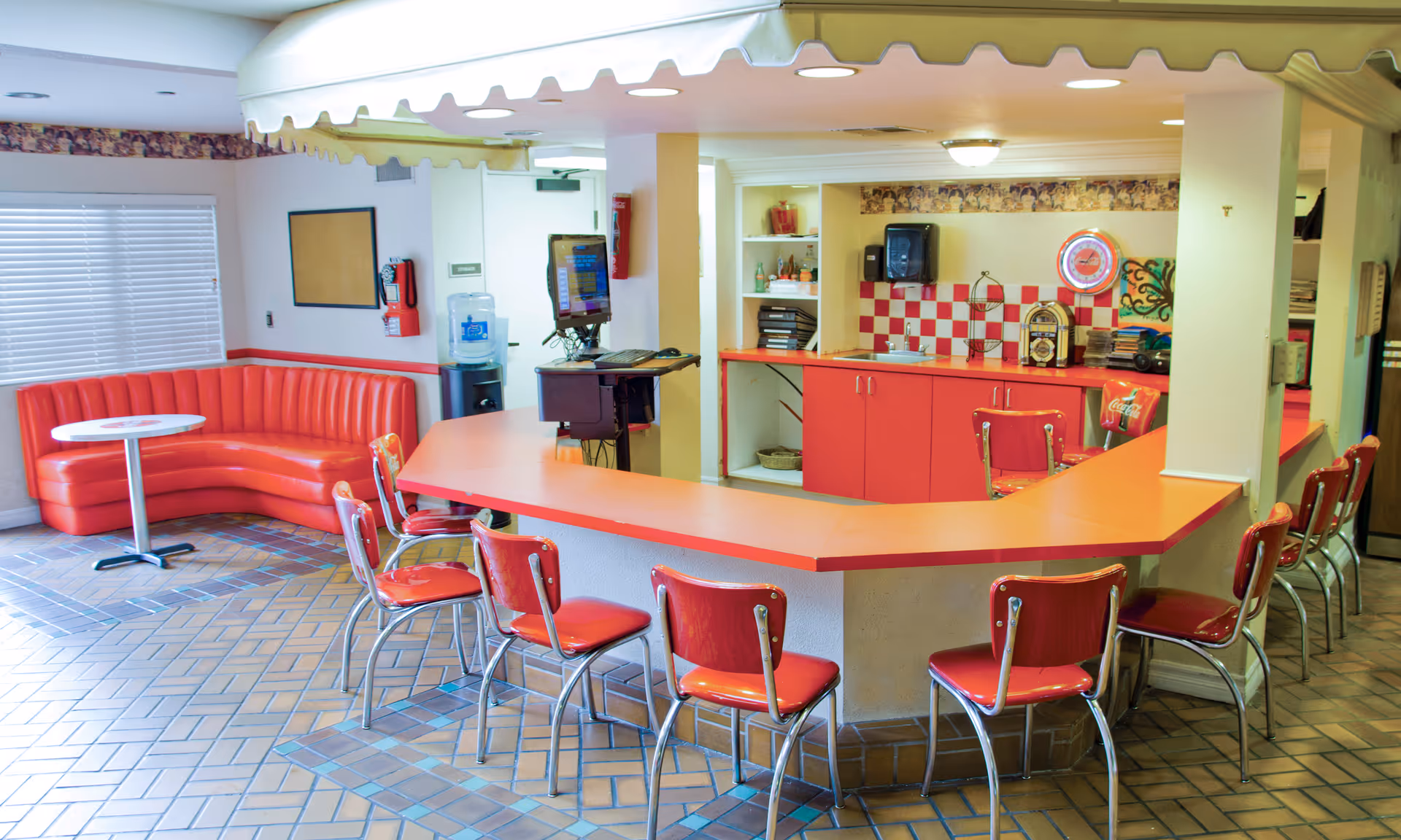 Interior view of a retro-style dining or lounge area with a red counter and matching red chairs arranged around it. There is a red cushioned booth seating area with a small round table near a window with blinds. The counter area has a small sink, shelves with various items, and a Coca-Cola themed wall clock and chair. The floor is tiled with a patterned design.