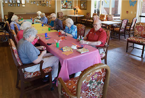 A group of elderly people sitting around a long table covered with colorful tablecloths in a well-lit room with wooden floors and floral upholstered chairs. They appear to be engaged in a social or recreational activity, with various items like cups and papers on the table. The room has large windows and additional tables and chairs in the background.