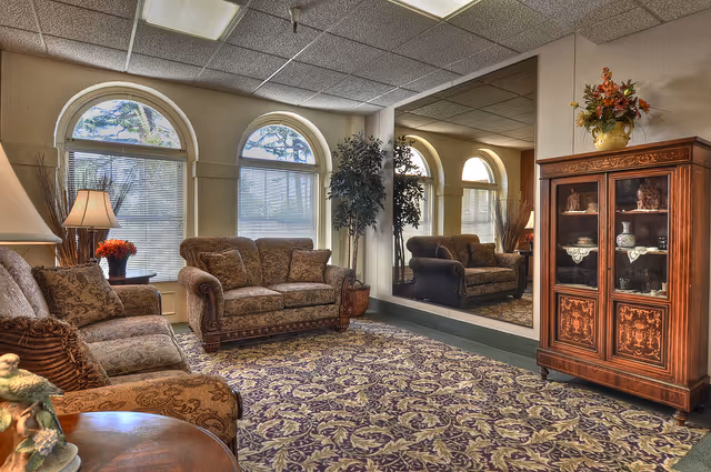 A cozy living room with patterned sofas and armchairs arranged around a large floral carpet. There are three arched windows with blinds, a tall potted plant, a wooden cabinet with decorative items, and a large mirror on one wall reflecting the room. A table lamp and a vase with flowers add warmth to the space.