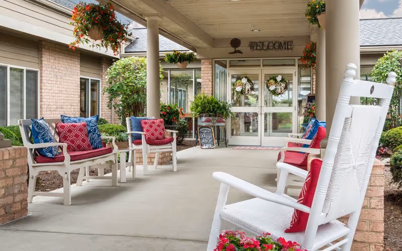 Covered outdoor seating area at Bickford of Middletown with white rocking chairs and benches adorned with red and blue cushions. Hanging flower baskets and potted plants decorate the space. Glass double doors with welcome signs lead into the building.