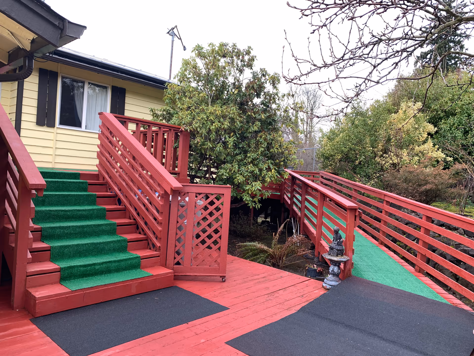 Outdoor view of a red wooden deck with green carpeted stairs and a ramp leading to a yellow building with black window shutters. There are trees and bushes surrounding the deck area.