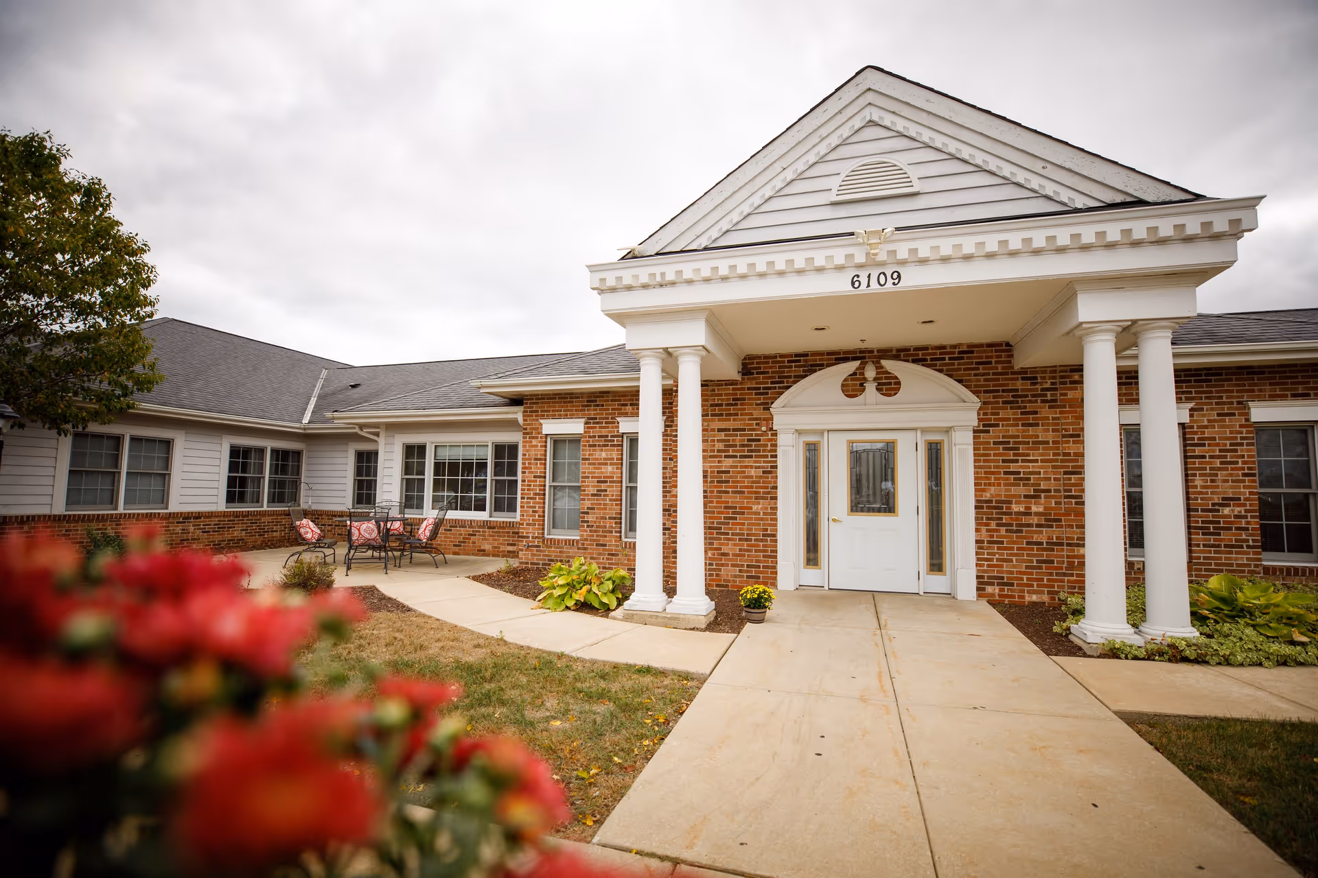 Front exterior view of a brick and white siding building with a covered entrance supported by four white columns. The entrance has a white door with glass panels and the number 6109 above it. There is a concrete walkway leading to the door, some landscaping with green plants and flowers, and a small patio area with a table and chairs with red cushions. The sky is cloudy.