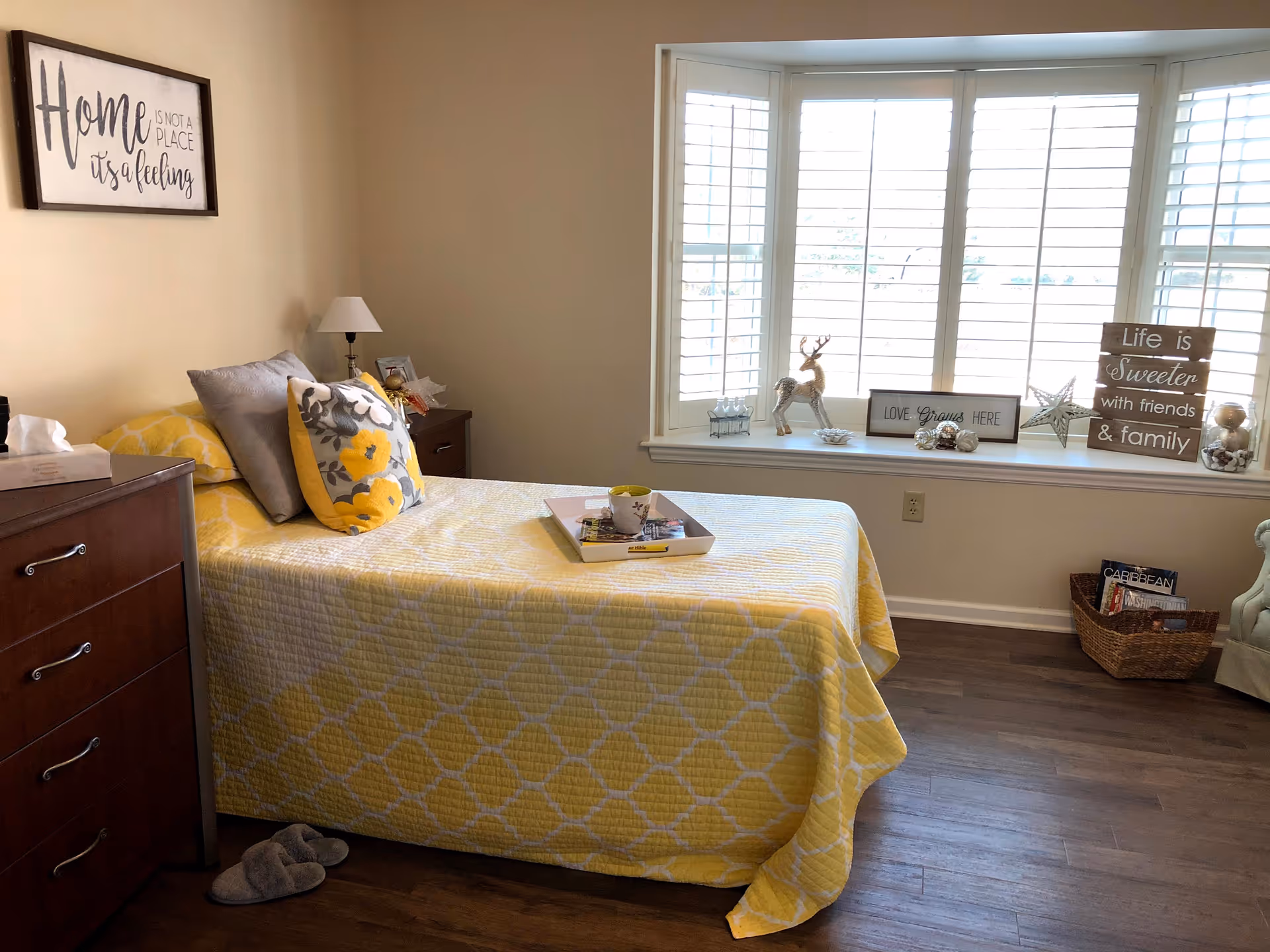 A cozy bedroom with a single bed covered in a yellow patterned quilt and decorative pillows. A wooden dresser with a tissue box and a bedside table with a lamp are visible. The room has a large window with white shutters and a window seat decorated with various ornaments and signs, including one that says 'Life is Sweeter with friends & family'. A pair of slippers is on the wooden floor next to the bed.