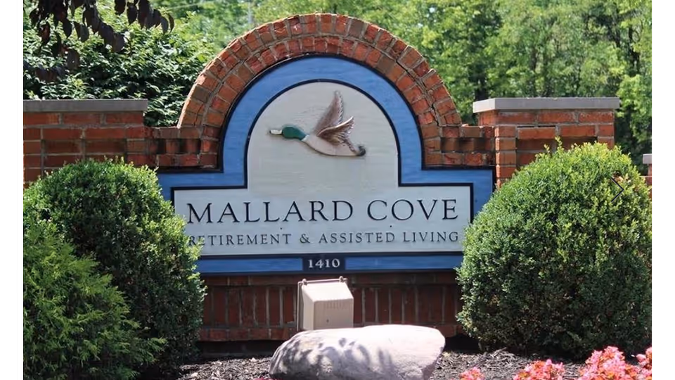 Brick sign with a blue and white background featuring a flying mallard duck illustration and the text 'Mallard Cove Retirement & Assisted Living 1410', surrounded by green bushes and flowers.