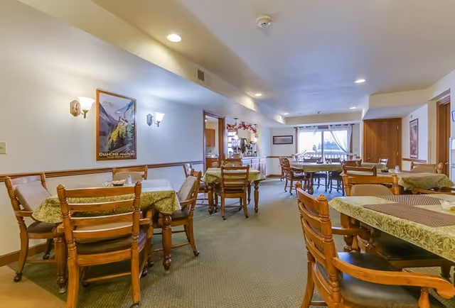A dining room in a senior living facility with several wooden tables and chairs covered with green tablecloths. The room is well-lit with wall sconces and ceiling lights, and there is a large window with curtains at the far end letting in natural light. A framed poster hangs on the wall, and the room has a cozy, inviting atmosphere.