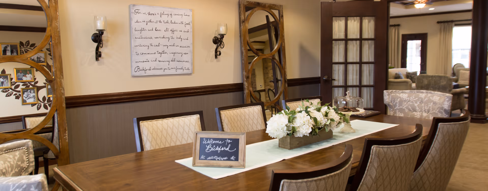 A cozy dining room with a long wooden table, upholstered chairs, a floral centerpiece and a small 'Welcome to Bickford' sign, with seating and decor visible beyond glass doors.