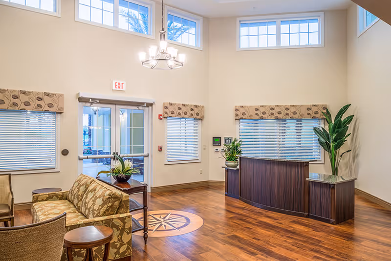 A bright and spacious assisted living facility lobby with wooden flooring, a decorative compass design on the floor, a wooden reception desk with plants on either side, a floral patterned sofa, chairs, and windows with blinds and valances. There is a chandelier hanging from the ceiling and an exit door with glass panels.