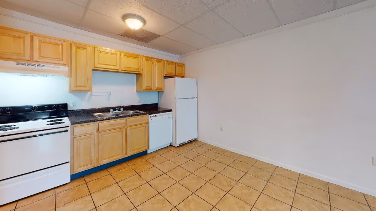 A kitchen area with light wood cabinets, a white refrigerator, a white dishwasher, a white electric stove with a range hood, a double sink, and tiled floor. The walls are plain white and the ceiling has recessed lighting.