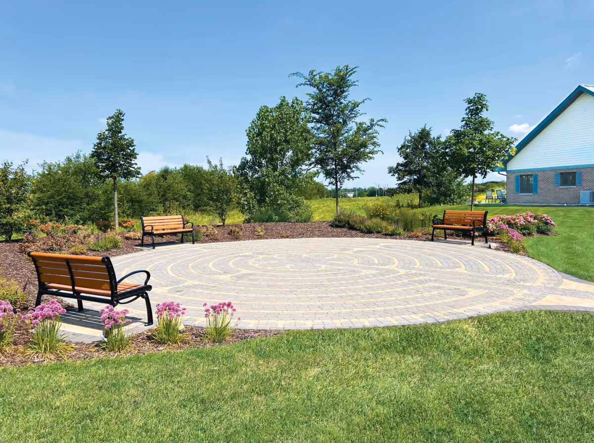 Outdoor circular paved area with a labyrinth design, surrounded by three wooden benches with black metal frames, green grass, flower beds with pink flowers, and several trees. A building with a white and brick exterior is visible in the background under a clear blue sky.