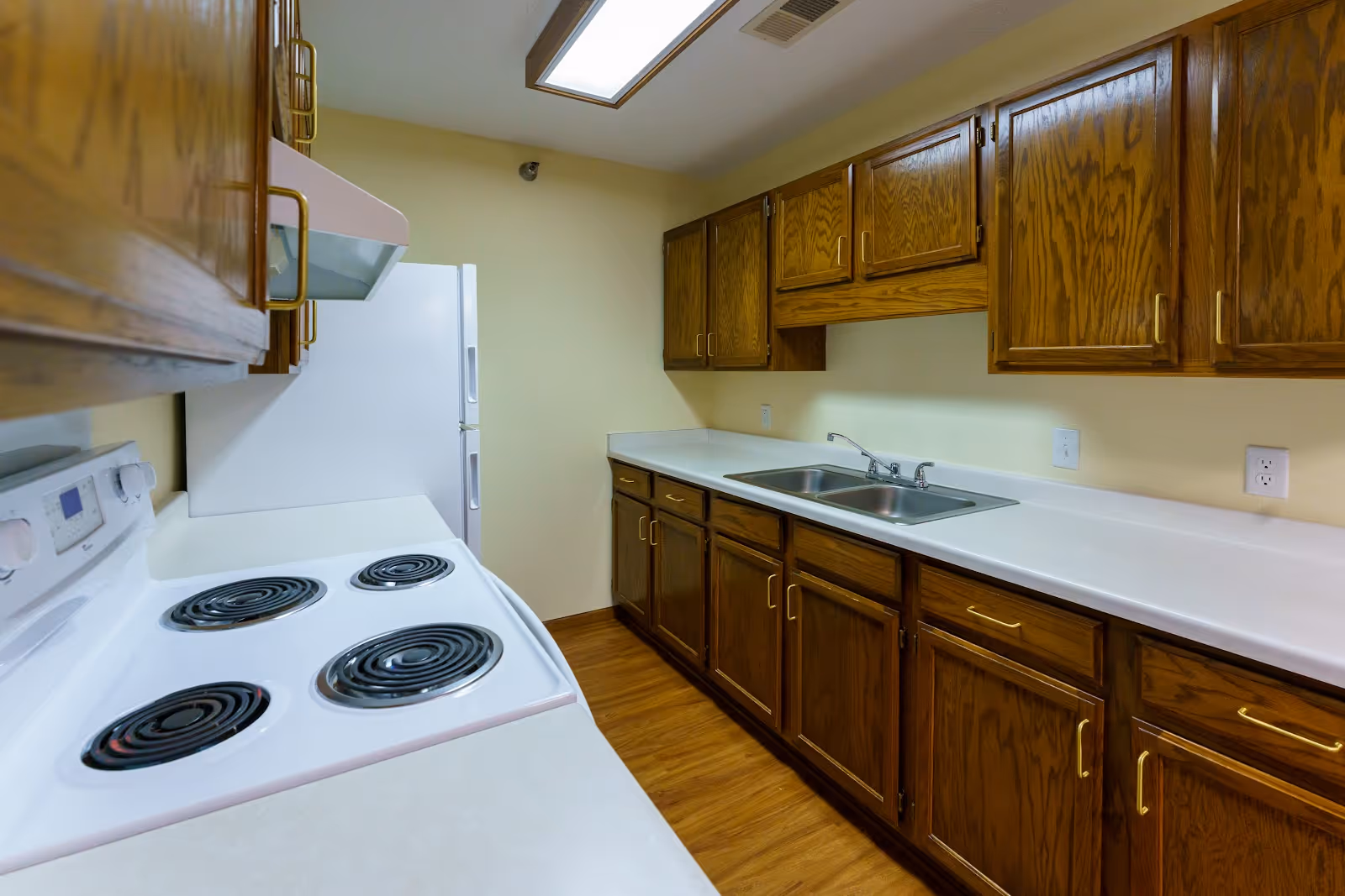 Interior view of a kitchen with wooden cabinets, a white electric stove with coil burners, a white refrigerator, a double sink, and white countertops under fluorescent ceiling lighting.