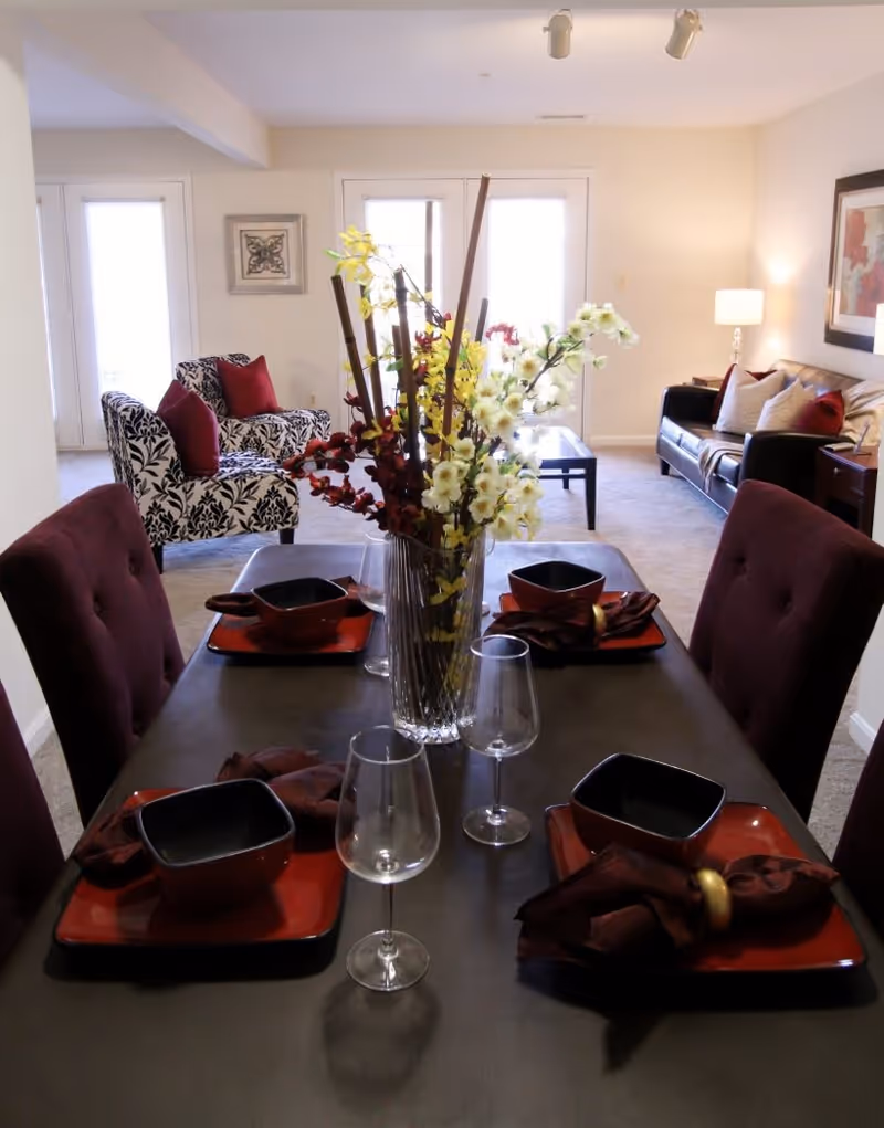 Dining table set with red and black place settings and a tall floral centerpiece in an open living room area.